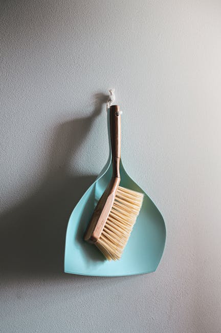 A light blue dustpan and a wooden-handled brush with natural bristles hanging on a plain, smooth, light grey wall, illuminated by soft, natural lighting to highlight their clean and well-maintained su