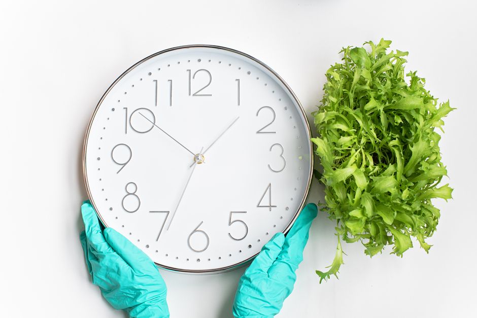 A person wearing bright blue rubber gloves is carefully holding a large, round wall clock with a silver frame, white face, and black numerals and hands, showing the time at approximately 1:48. The clo